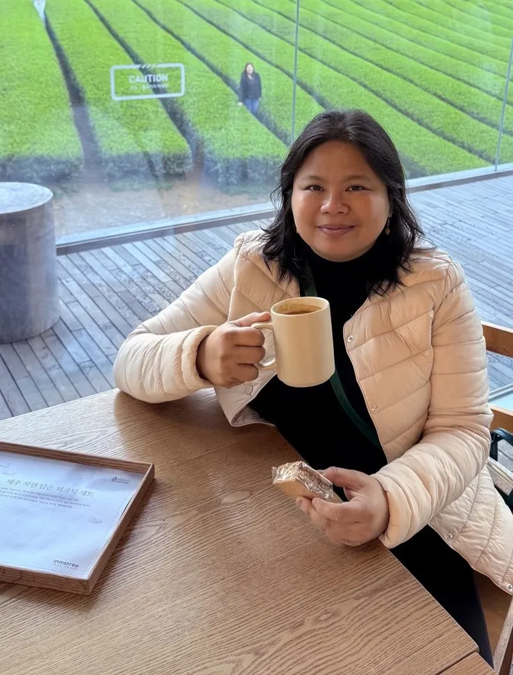 Rea Celine Villa holding a mug at a wooden table by a window